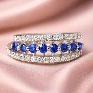 May include: A silver ring with a double band design. The top band is encrusted with small, clear gemstones. The lower band features a row of vibrant blue gemstones. The ring is set against a soft pink background.