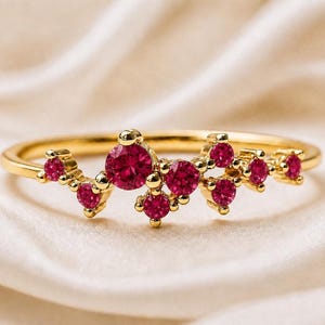 Könnte beinhalten: Ein goldener Ring mit einer Ansammlung leuchtend roter Edelsteine. Der Ring hat ein schmales, poliertes Goldband, und die Edelsteine sind in einem zarten, asymmetrischen Muster angeordnet. Der Ring liegt auf einem weichen, cremefarbenen Hintergrund.