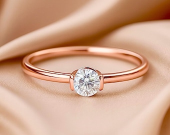 May include: Rose gold ring with a round, clear gemstone. The ring is set on a soft, peach-colored fabric. The band is thin and delicate, with a simple, elegant design. The gemstone is the focal point, catching the light.
