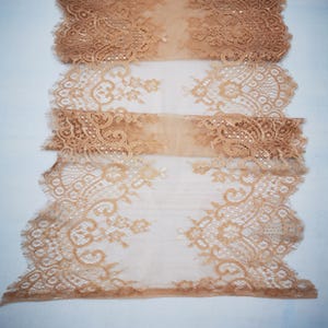 May include: A tan lace table runner with intricate floral and geometric patterns. The runner features a delicate, scalloped edge and is made of a sheer, lightweight fabric. The design is visible against a white background.