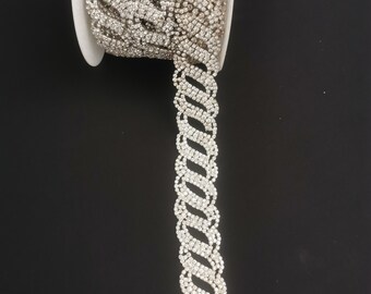 May include: A roll of silver rhinestone trim with a repeating chain-like design. The trim is on a white spool against a black background. The trim is made of many small, sparkling rhinestones.