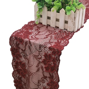 May include: A burgundy lace table runner featuring a floral design. The runner is displayed with a small white picket fence and greenery. The lace has an intricate pattern, adding a touch of elegance to any table setting.