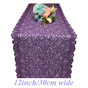 Könnte beinhalten: Lilafarbener Tischläufer aus Spitze mit einem zarten Blumenmuster. Der Läufer ist 30 cm breit und hat einen gewellten Rand.