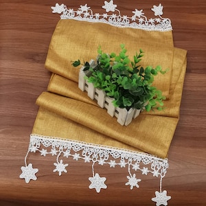 May include: A gold table runner with white lace trim and star-shaped fringe. The runner is folded and a small potted plant is sitting on top of it.