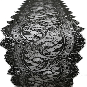 May include: Black lace table runner featuring a detailed floral and paisley pattern. The runner has a scalloped edge and is made from a delicate, see-through material. The design repeats along the length, creating an elegant aesthetic for any table setting.