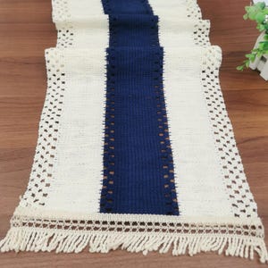 May include: White table runner with a blue stripe down the center. The runner has a crocheted border and fringe.