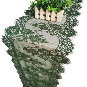 Può includere: Un runner da tavolo in pizzo verde con un delicato motivo floreale. Il runner è sfrangiato sui bordi e presenta un design a festoni. Una piccola pianta in vaso è appoggiata sul runner.