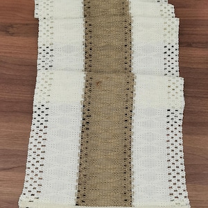 May include: A white crocheted table runner with a brown woven stripe down the center. The runner has a fringe border.
