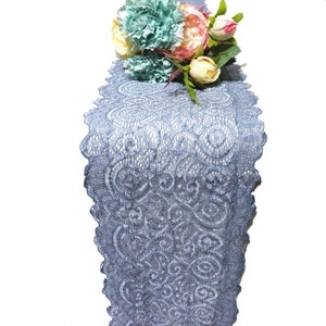 Könnte beinhalten: Ein blauer Tischläufer aus Spitze mit einem zarten Blumenmuster. Der Läufer ist mit einem Strauß künstlicher Blumen in den Farben Blau, Rosa und Weiß verziert.
