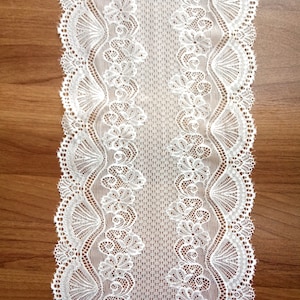 May include: White lace trim with a delicate floral pattern. The lace is on a brown wooden surface.