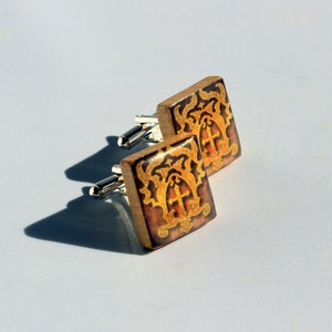 May include: A pair of square cufflinks with a wooden base and silver stems. The cufflinks have a brown background with a gold and red ornate design. The design includes a cross and decorative flourishes.