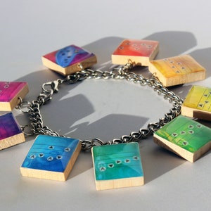 May include: A silver charm bracelet featuring nine square charms with colorful, abstract designs. Each charm is a different color, including blue, green, yellow, and pink, with a wooden base and a glossy finish. The charms have a musical theme.