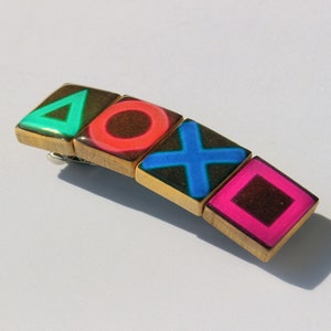 May include: A gold hair clip with four square tiles featuring the PlayStation controller symbols: a green triangle, a red circle, a blue X, and a pink square.