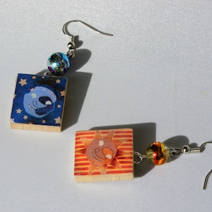 Puede incluir: Un par de pendientes con un diseño de calavera azul y blanco y un diseño de sol rojo y naranja. Los pendientes están hechos de madera y tienen un gancho de metal.