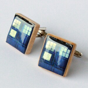 May include: A pair of cufflinks featuring a blue and silver image of the TARDIS, the time machine from the Doctor Who television series. The cufflinks are made of wood and have a silver metal clasp.