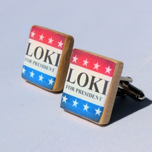 May include: A pair of wooden cufflinks with a red, white and blue American flag design. The cufflinks feature the text "Loki for President".