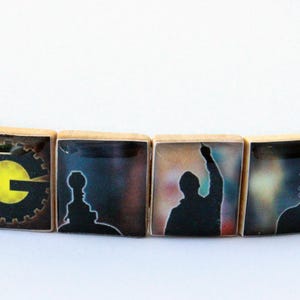 May include: A wooden bracelet with four square beads. The first bead has a yellow "G" inside a black gear. The other beads have silhouettes of figures against colorful backgrounds. The bracelet is on a white background.
