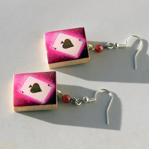 May include: A pair of earrings featuring a pink and white playing card with a black spade. The card is attached to a silver hook earring.