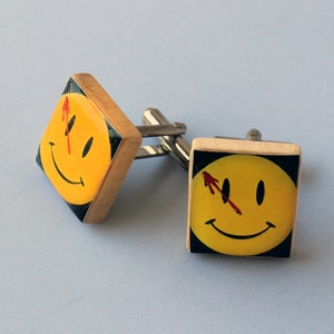 May include: A pair of cufflinks featuring a yellow smiley face with a red slash through the eye. The cufflinks are made of wood and have a silver metal connector.
