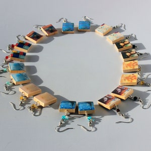 May include: A collection of 18 square wooden earrings with various colorful images printed on them. The earrings are arranged in a circle on a white surface.
