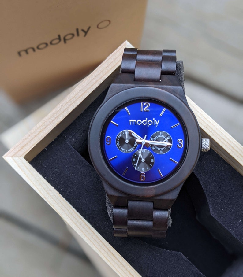 Op de afbeelding: Een donkerbruin houten polshorloge met een blauwe wijzerplaat. Het horloge heeft een chronograaf en een dag van de week weergave. Op de wijzerplaat van het horloge staat "modply".
