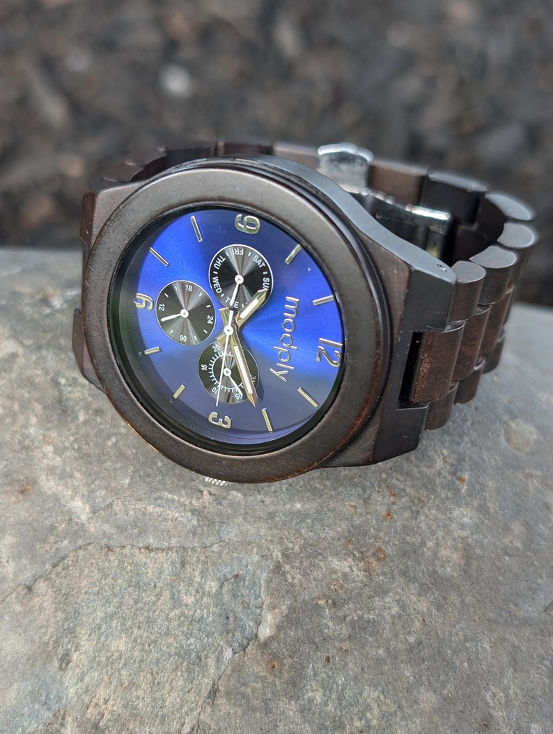 Op de afbeelding: Een donkerbruin houten polshorloge met een blauwe wijzerplaat en gouden accenten. De wijzerplaat van het horloge heeft een dag van de week en datumweergave. Het horlogemerk is "modaly".