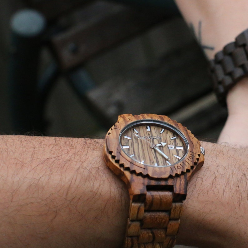 May include: A wooden wristwatch with a brown and black wood grain pattern. The watch face has a white background with black markings and the word "woodply" printed on it.