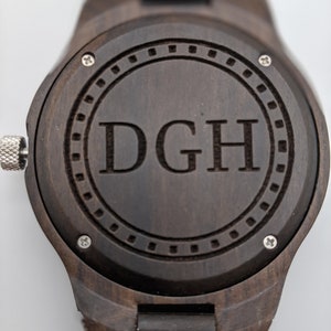 Op de afbeelding: Een donkerbruin houten polshorloge met een ronde wijzerplaat. De wijzerplaat heeft een zilveren ring rond de rand met een cirkel in het midden. De initialen "DGH" zijn gegraveerd in de middelste cirkel.