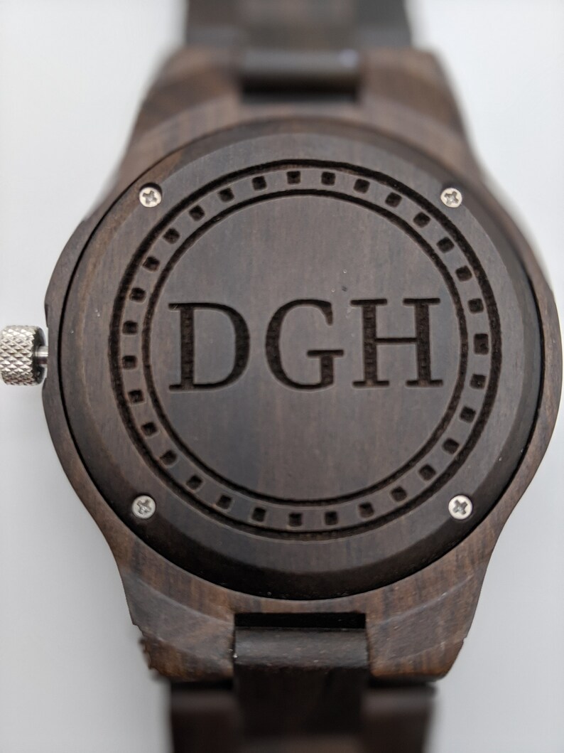 Op de afbeelding: Een donkerbruin houten polshorloge met een ronde wijzerplaat. De wijzerplaat heeft een zilveren ring rond de rand met een cirkel in het midden. De initialen "DGH" zijn gegraveerd in de middelste cirkel.