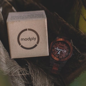 May include: A wooden watch with a dark brown face and matching band sits next to a cardboard box. The watch face has white hands and the brand name "modply" is printed on the box and watch face.