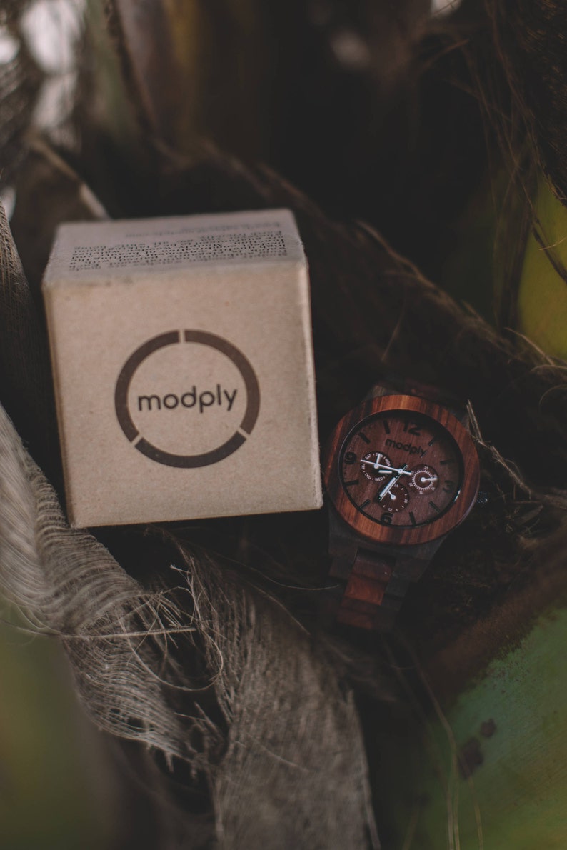 May include: A wooden watch with a dark brown face and matching band sits next to a cardboard box. The watch face has white hands and the brand name "modply" is printed on the box and watch face.