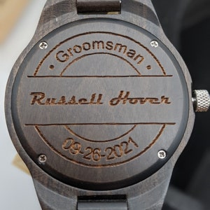 Op de afbeelding: Een donkerbruin houten polshorloge met een ronde wijzerplaat. De wijzerplaat is gegraveerd met de tekst "Groomman", "Russell Hoover" en "09-26-2021".