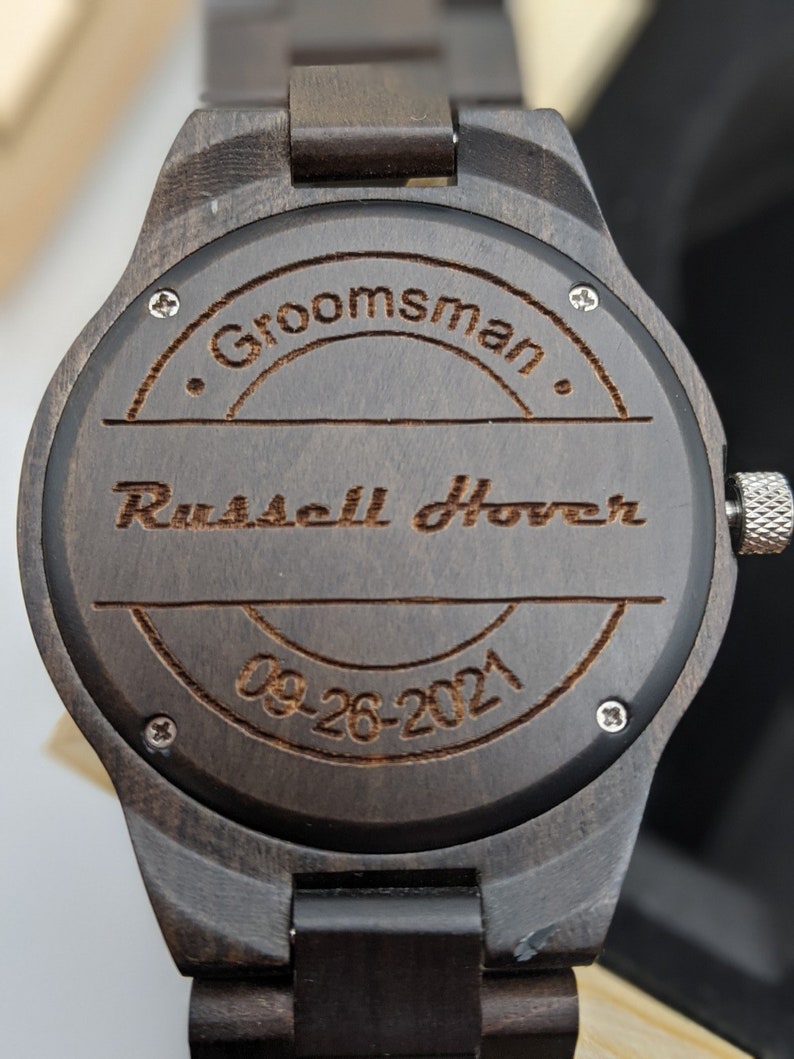 Op de afbeelding: Een donkerbruin houten polshorloge met een ronde wijzerplaat. De wijzerplaat is gegraveerd met de tekst "Groomman", "Russell Hoover" en "09-26-2021".