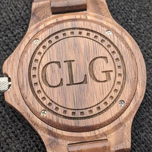 May include: A brown wooden wristwatch with a dark brown band and a circular face. The face has a concentric circle design with the initials "CLG" engraved in the centre.