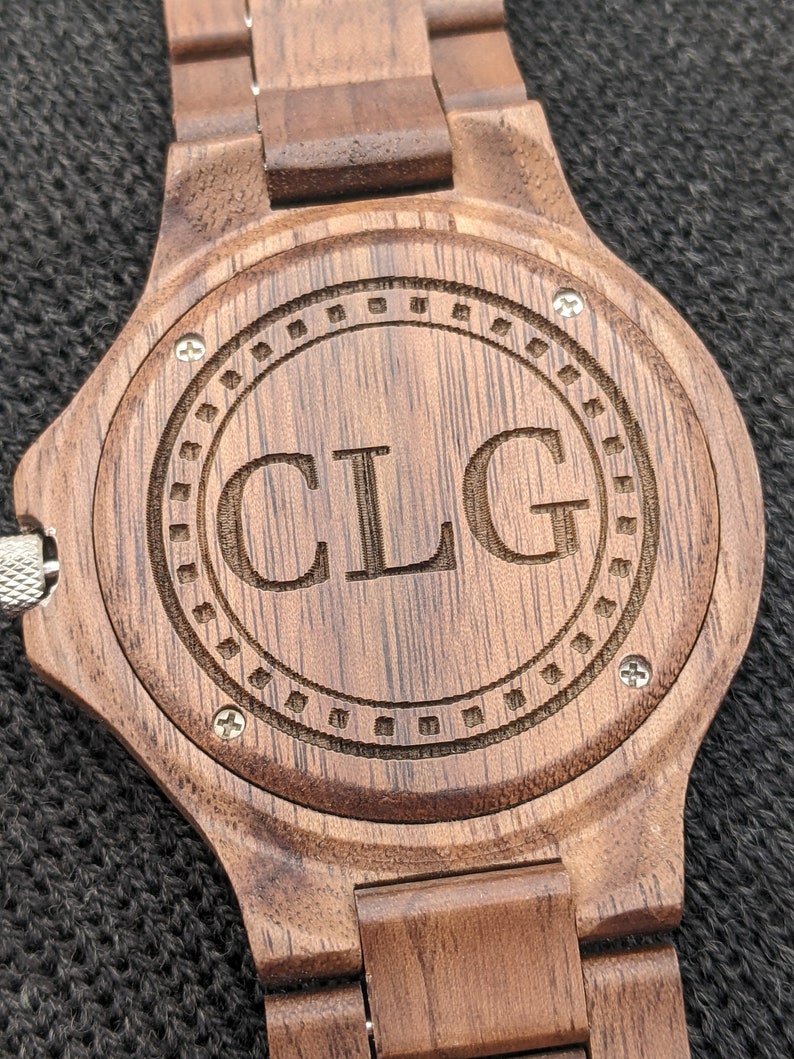 May include: A brown wooden wristwatch with a dark brown band and a circular face. The face has a concentric circle design with the initials "CLG" engraved in the centre.