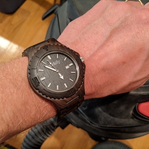 May include: A dark brown wooden wristwatch with a black face and white markings. The watch has a black leather band and a silver buckle.