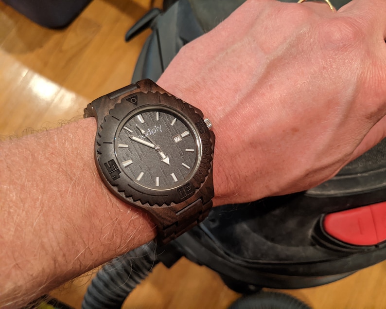 May include: A dark brown wooden wristwatch with a black face and white markings. The watch has a black leather band and a silver buckle.
