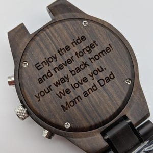 Op de afbeelding: Een donkerbruin houten polshorloge met een zwarte band. De achterkant van het horloge is gegraveerd met de boodschap "Enjoy the ride and never forget your way back home! We love you, Mom and Dad."