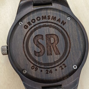 Op de afbeelding: Een donkerbruin houten horloge met een zwarte wijzerplaat en gegraveerde tekst die "Groomsman" en "SR 09.24.22" luidt.
