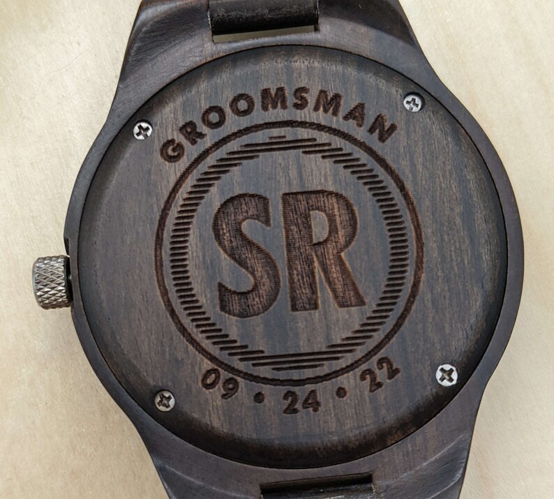 Op de afbeelding: Een donkerbruin houten horloge met een zwarte wijzerplaat en gegraveerde tekst die "Groomsman" en "SR 09.24.22" luidt.