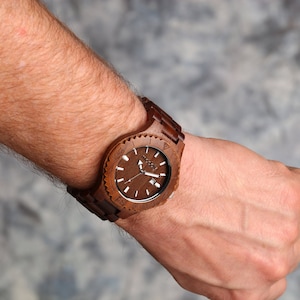 May include: A brown wooden wristwatch with a black face and white markings. The watch has a unique design with a wooden band and a circular face.