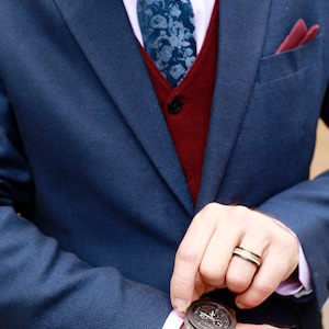 May include: A navy blue suit with a burgundy vest and a floral patterned tie. A wooden watch with a dark face and band is visible. A ring with black, gold, and silver bands is also visible. The suit has a pink shirt and a burgundy pocket square.