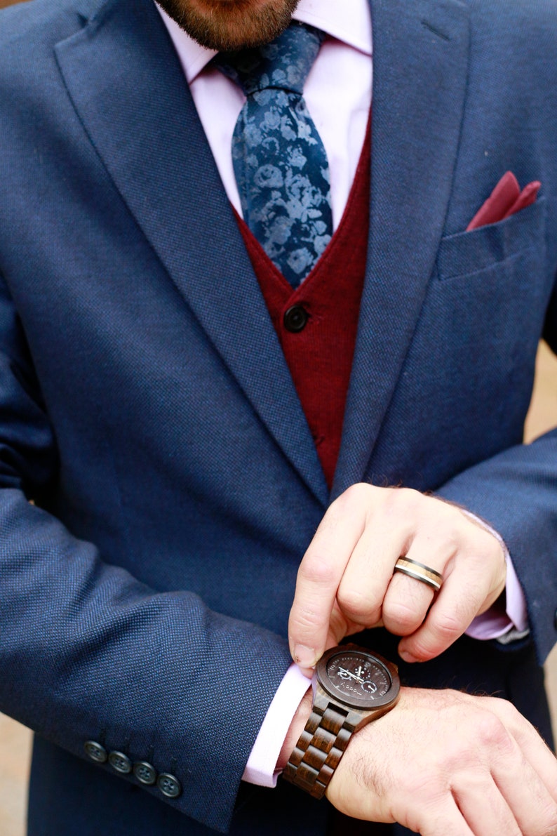 May include: A navy blue suit with a burgundy vest and a floral patterned tie. A wooden watch with a dark face and band is visible. A ring with black, gold, and silver bands is also visible. The suit has a pink shirt and a burgundy pocket square.
