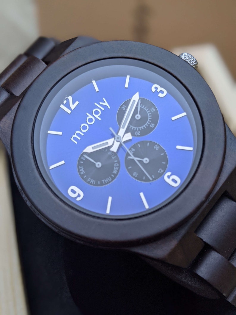 Op de afbeelding: Een donkerbruin houten polshorloge met een blauwe wijzerplaat. Het horloge heeft drie subwijzerplaten en het woord "modply" staat op de wijzerplaat gedrukt.