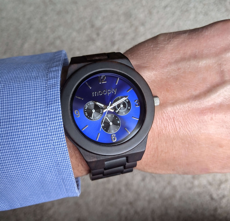 Op de afbeelding: Een zwart houten polshorloge met een blauwe wijzerplaat en zilveren wijzers. Het horloge heeft een datumvenster en een dag van de week venster. Op de wijzerplaat van het horloge staat ook het woord "moodply" gedrukt.