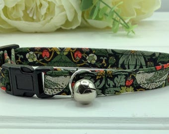 May include: A patterned cat collar with a black plastic buckle and a silver bell. The fabric features a dark green background with a floral and bird design in red, white, and yellow.