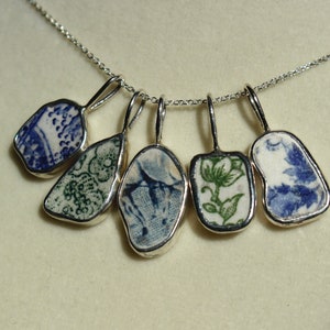 Könnte beinhalten: Eine silberne Halskette mit fünf Anhängern, die jeweils ein anderes blaues und grünes Blumenmuster auf weißem Hintergrund zeigen.