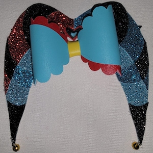 May include: A whimsical jester hat with a large, light blue bow and red scalloped edges. The hat features alternating sections of black, red, and blue glitter, with small gold bells at the tips. A yellow band connects the bow.