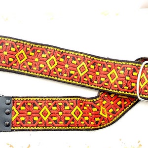 Op de afbeelding: Een rode, gele en zwarte gitaarband met een geometrisch patroon en een zilveren gesp.