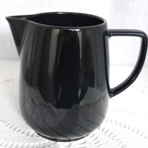 Puede incluir: Una jarra de crema de cerámica negra con un acabado liso y brillante y un cuerpo redondeado. La jarra tiene una sola asa.